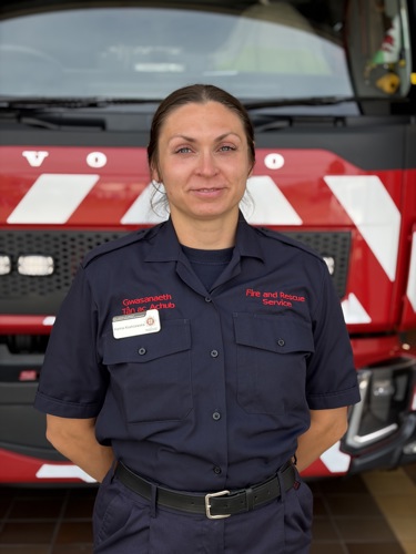 Photo of a woman in Fire and Rescue Service uniform standing in front of a red fire engine with arms behind her back.